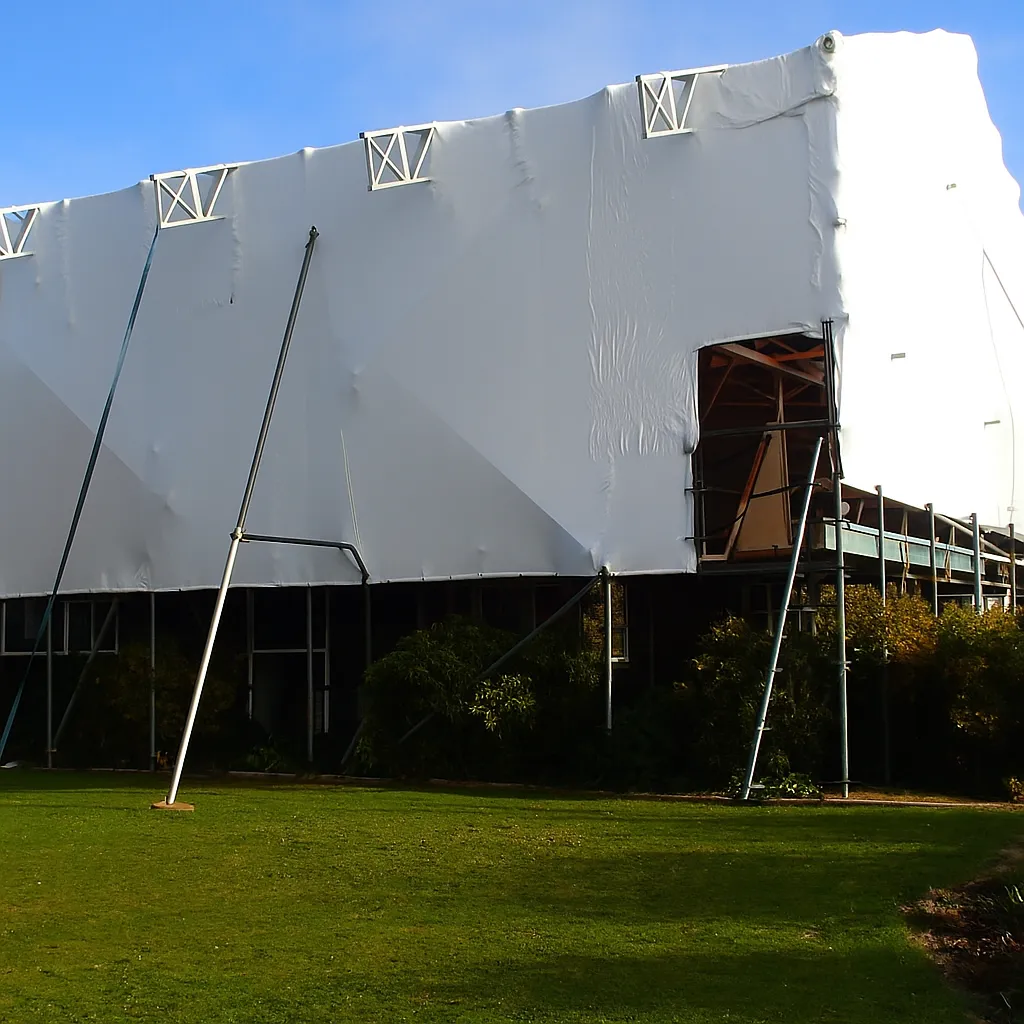 A residential building under repair is fully covered in white shrink wrap with scaffolding and support beams visible.