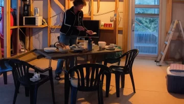 live in house during renovation. A woman preparing food at a makeshift dining table inside a partially renovated home, surrounded by plastic chairs, construction materials, and exposed framing.
