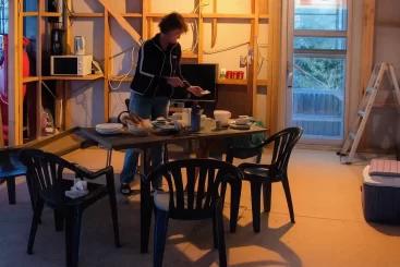 live in house during renovation. A woman preparing food at a makeshift dining table inside a partially renovated home, surrounded by plastic chairs, construction materials, and exposed framing.