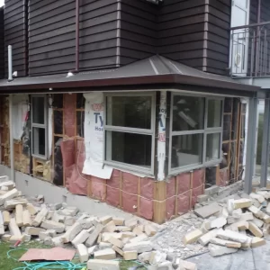 Exterior view of a residential building mid-renovation, with cladding removed and insulation exposed, surrounded by rubble and loose bricks.