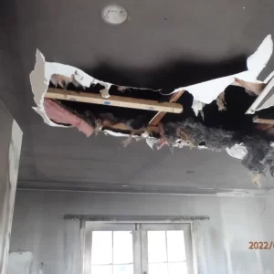 Interior ceiling of a smoke-damaged room with a large hole exposing charred insulation and wooden framing, suggesting fire or structural damage.