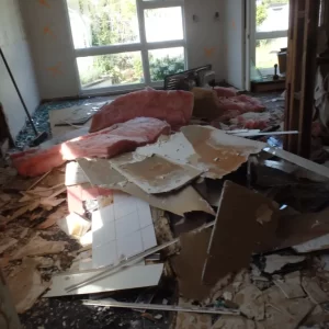 Interior room filled with building debris including broken plasterboard, tiles, insulation, and shattered glass, with orange markings on the walls and visible structural damage.