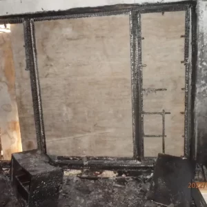 Charred interior wall and window frame after a fire, with extensive smoke and burn damage visible. Burnt debris and ash cover the floor.